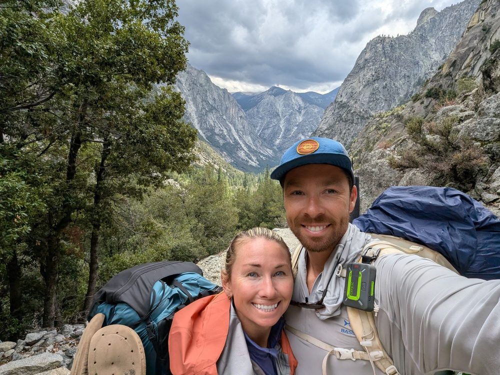 Chris and his wife Lauren smiling in hiking gear on a mountain trail, with a granite valley behind them